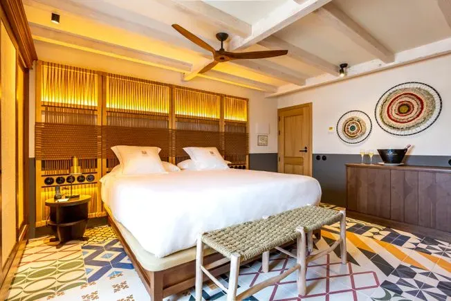 Dormitorio de hotel con cama tamaño king, cabecera tejida, banco y piso con estampado colorido.