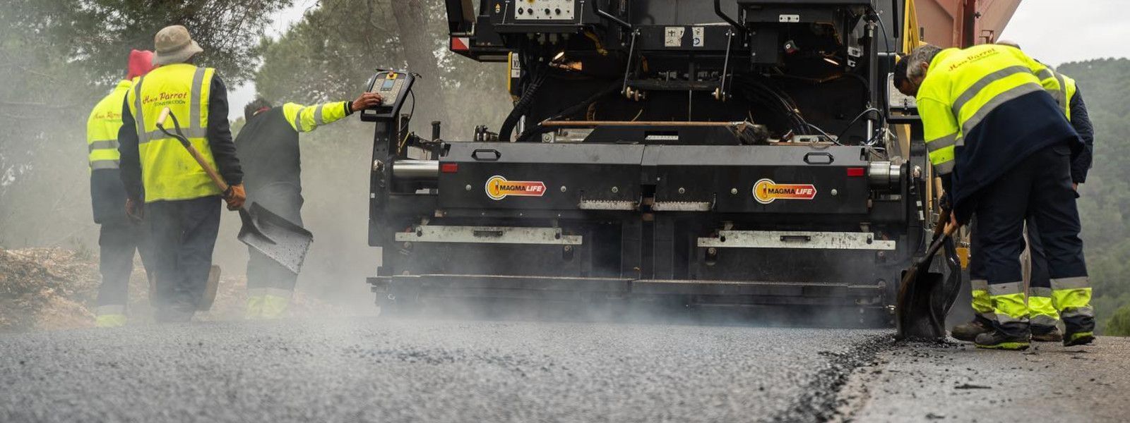 Trabajadores de la carretera pavimentando una carretera con una máquina, vistiendo chalecos de seguridad de color amarillo brillante.