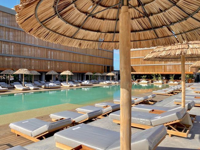 Escena junto a la piscina con sombrillas de paja, tumbonas y una piscina turquesa. Edificios al fondo.