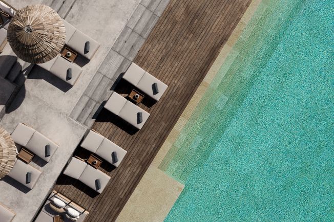 Tumbonas con sombrillas de paja junto a una piscina turquesa. Terraza de madera y zona de arena.