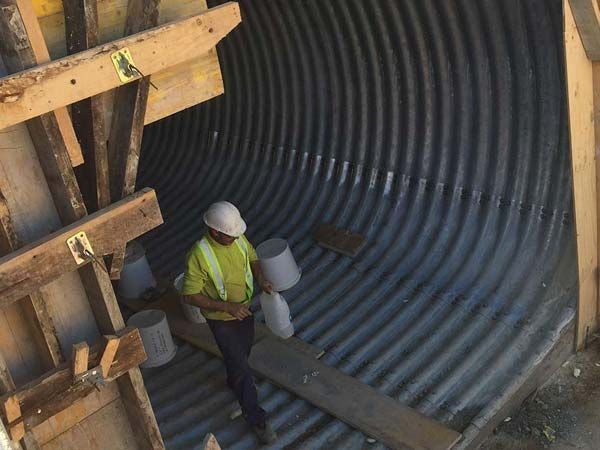 Un hombre con casco de seguridad dentro de un túnel de metal corrugado, sosteniendo dos cubos. Soportes de madera enmarcan la abertura.