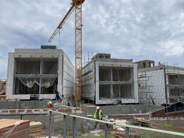 Obra en construcción con estructuras de hormigón en forma de caja; grúa; trabajadores con chalecos, cielo nublado.