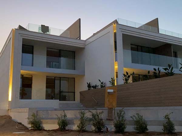 Moderna casa blanca de dos pisos con balcones de vidrio, cerca marrón e iluminación exterior.