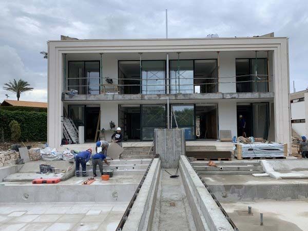 Obreros de la construcción alrededor de un edificio de dos plantas con amplios ventanales y dos piscinas en construcción. Día nublado.