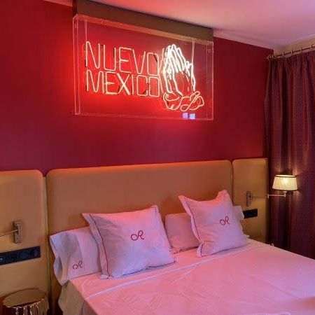 Recámara con paredes rojas, cabecera amarilla, cama con sábanas blancas y letrero de neón que dice