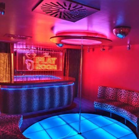 Interior de una discoteca con pista de baile, barra, tubo y luces de neón en rojo y azul.