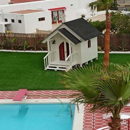 Pequeña casa blanca con una puerta roja cerca de una piscina, al lado de un césped verde y una palmera.