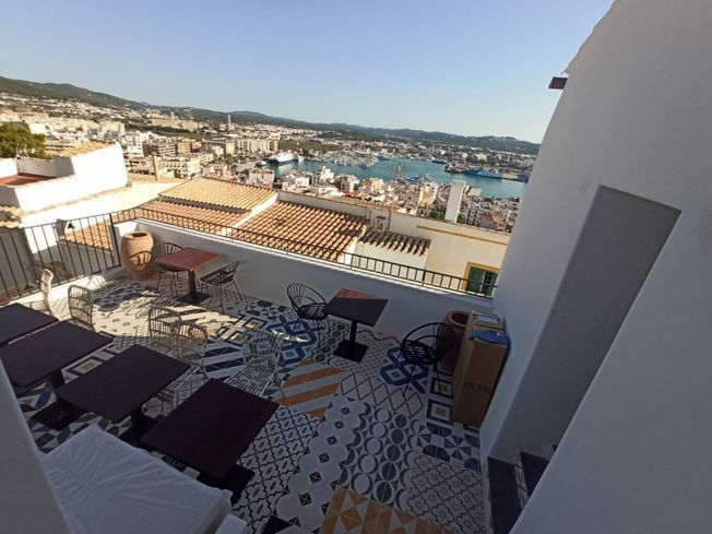 Restaurante en la azotea con vista al puerto, mesas, suelo de baldosas estampadas, cielo azul, edificio blanco.