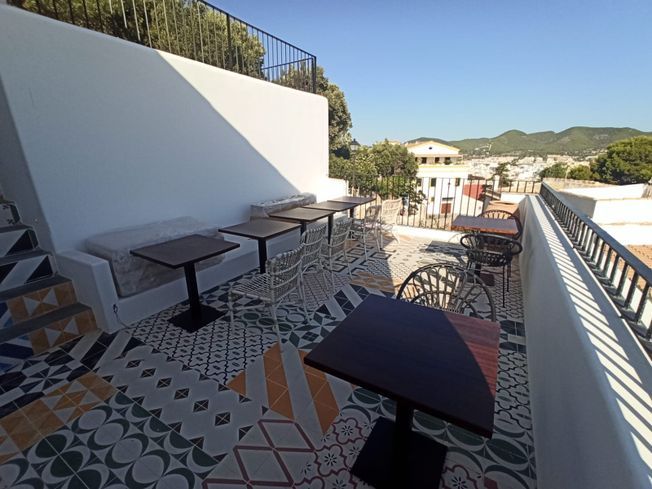 Terraza de cafetería al aire libre con mesas, azulejos estampados, paredes blancas, vista a la ciudad, día soleado.