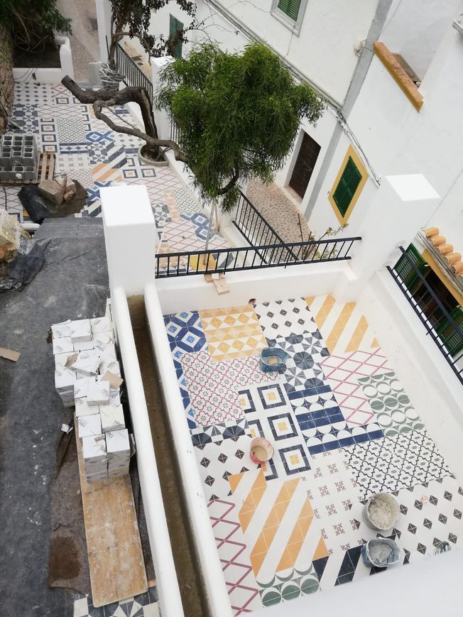 Un patio en la azotea con un colorido suelo de baldosas en construcción. También se ven edificios blancos, un pequeño árbol y una barandilla.