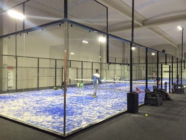 Pista de pádel con jugadores. Pista azul, paredes de cristal, estructura metálica, interior. Un jugador en acción.