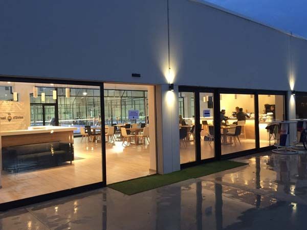 Exterior de una cafetería moderna al anochecer. Los ventanales permiten ver a los clientes sentados en las mesas. Las luces iluminan el exterior.
