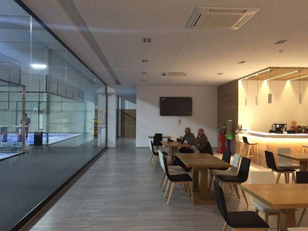 Interior de la cafetería de un club deportivo con vistas a una pista de pádel. Dispone de mesas, sillas y una zona de bar.
