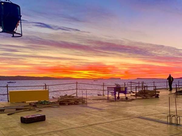 Atardecer sobre el agua desde una obra en construcción. Cielo naranja intenso y morado. Una figura solitaria camina.