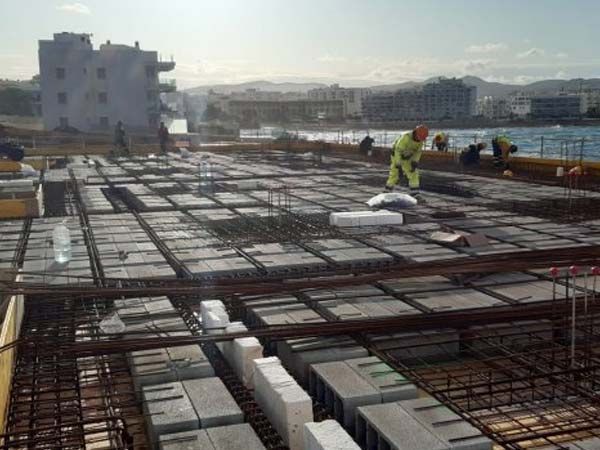 Obreros de la construcción sobre una losa de hormigón; varillas de refuerzo, bloques y equipo visibles. Al aire libre, cerca del agua, soleado.