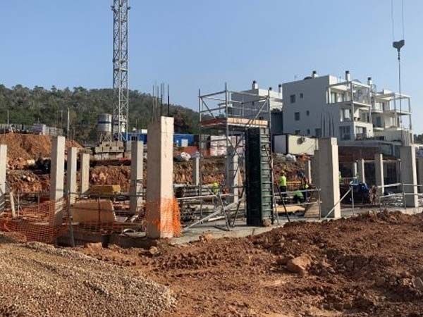 Sitio de construcción con columnas de hormigón, andamios y un edificio parcialmente construido bajo un cielo despejado.