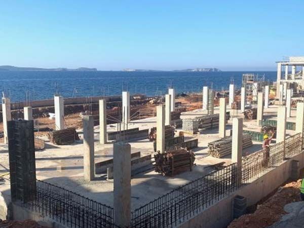 Sitio de construcción con pilares de hormigón, barras de refuerzo y fondo marino bajo un cielo azul.
