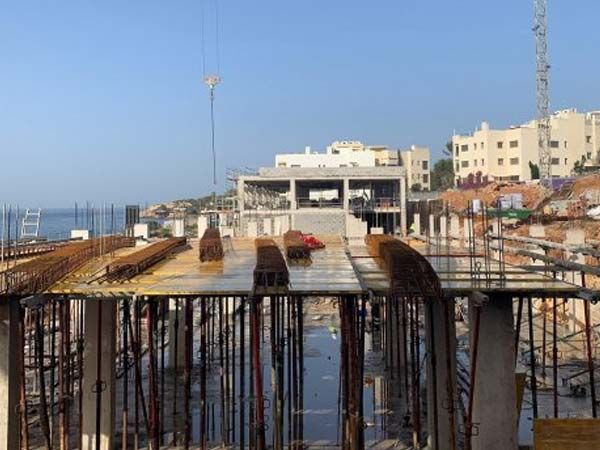 Sitio de construcción con varillas de refuerzo, encofrados de hormigón y un edificio parcialmente construido cerca de la costa.