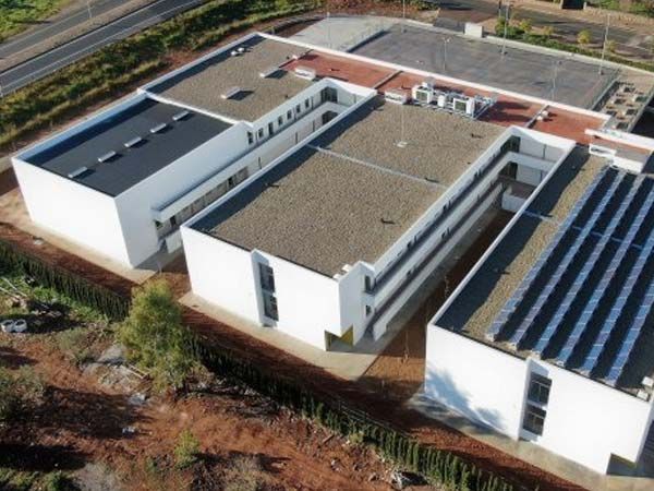 Vista aérea de un edificio moderno blanco con techos planos y paneles solares, al lado de una carretera y tierra firme.