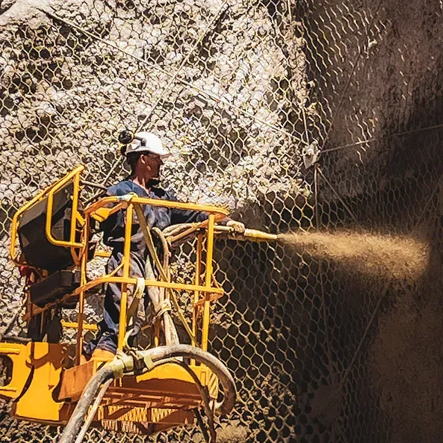 Un trabajador rocía hormigón sobre una pared rocosa, de pie en un elevador amarillo. Una malla cubre la roca, iluminada por el sol, al aire libre.