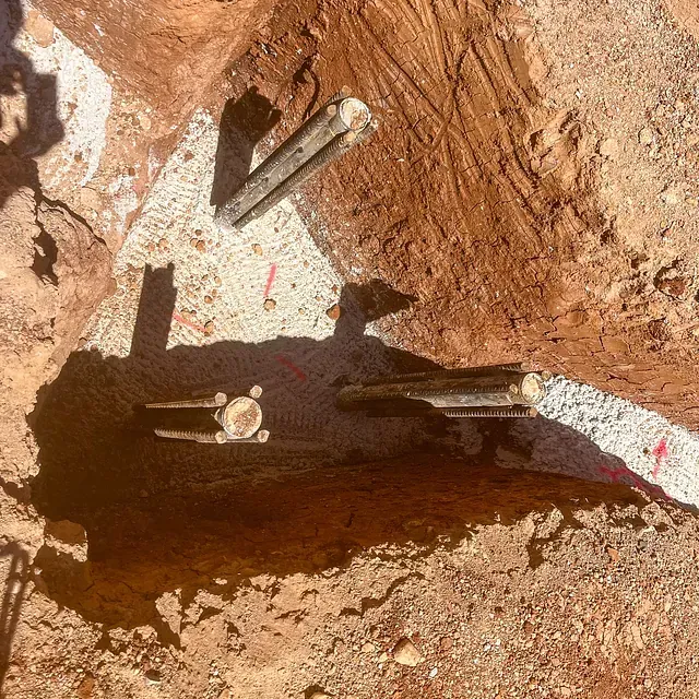 Vista aérea de un sitio de construcción con vigas de madera en agujeros excavados, rodeado de tierra y grava.