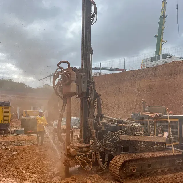 Sitio de construcción con plataforma de perforación, trabajador rociando agua, cielo nublado, suelo fangoso.