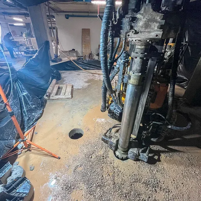 Una perforadora perfora un agujero en un suelo de hormigón. Obra con escombros y una persona al fondo.