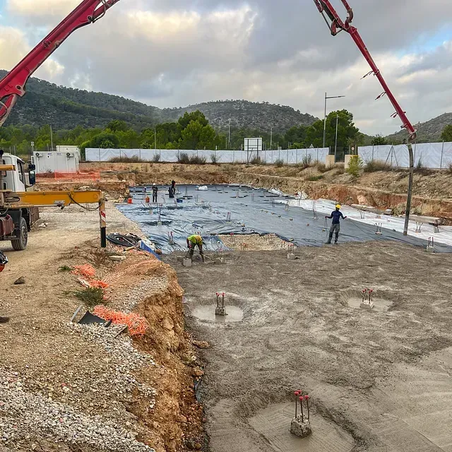 Sitio de construcción con hormigón siendo vertido; trabajadores, fondo de montaña.