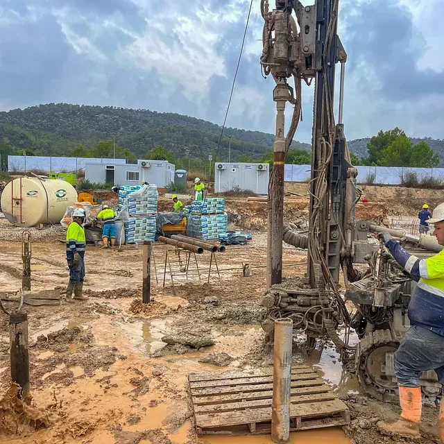 Obra en construcción. Se ven trabajadores con chalecos de seguridad, perforando, lodo, suministros y una perforadora.