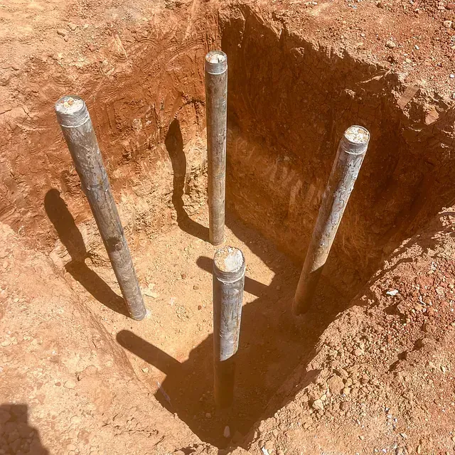 Cuatro postes cilíndricos en una excavación cuadrada. Suelo marrón rojizo. Día soleado.