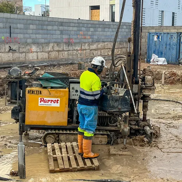 Trabajador de la construcción operando un taladro en un sitio fangoso, usando equipo de seguridad, con una máquina Parrot amarilla.