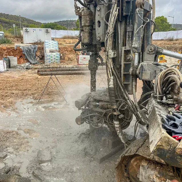 Máquina perforadora en funcionamiento, levantando barro y polvo en un sitio de construcción.