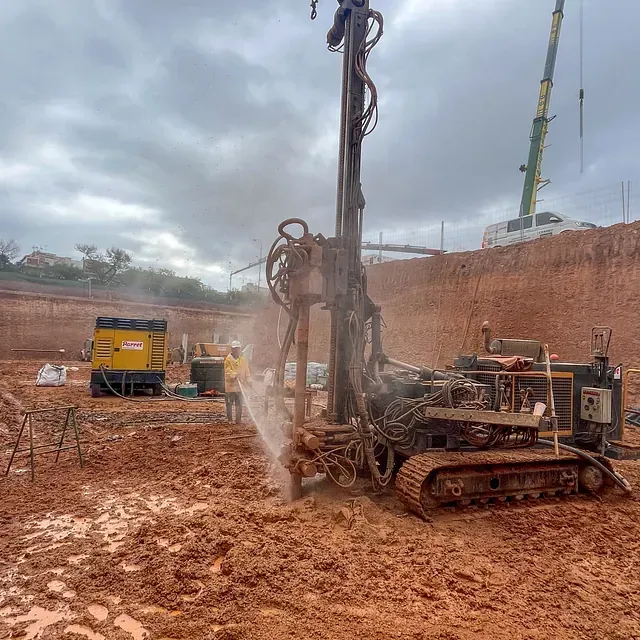 Sitio de construcción con máquina perforadora; trabajador rocía agua; cielo nublado; suelo fangoso.