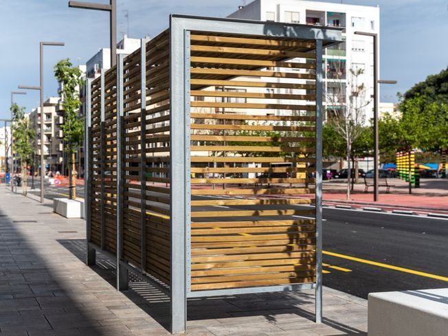 Estructuras de listones de madera sobre una acera, con marco de metal gris. Calle con edificios y árboles al fondo.