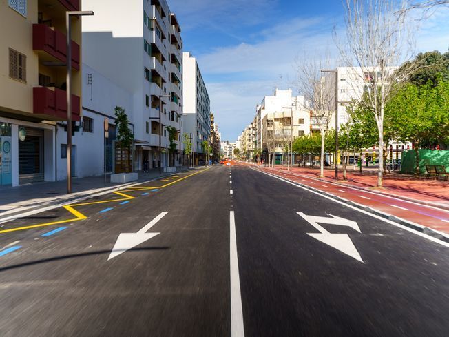 Calle con edificios blancos, árboles, carril bici rojo y cielo azul. Flechas en la carretera.