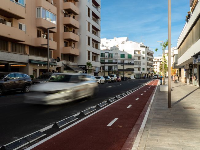 Escena callejera con carril bici. Un coche blanco en movimiento, rodeado de coches aparcados y edificios.