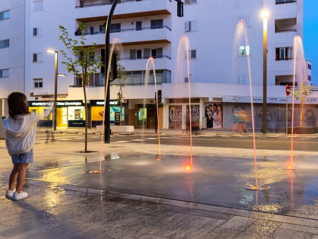 Niña contemplando las fuentes iluminadas de una plaza al anochecer. Edificios y farolas al fondo.