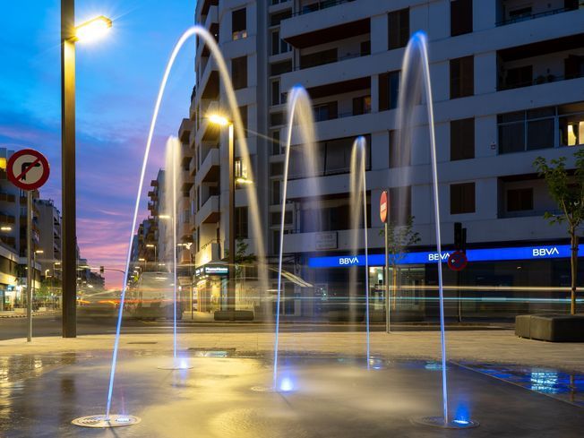 Fuentes que arrojan agua iluminadas en azul en una plaza al anochecer. Edificios y farolas al fondo.