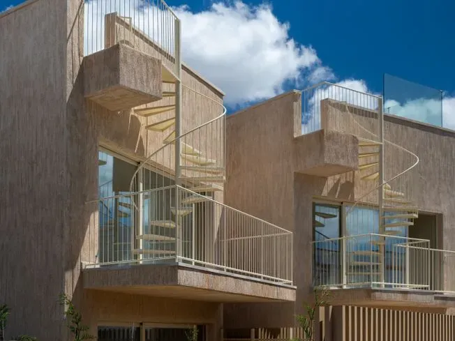 Edificios beige de dos pisos con barandillas de vidrio y escaleras de caracol, detrás de una valla de madera bajo un cielo azul.