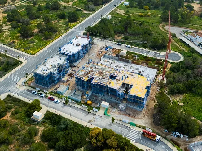 Vista aérea de un sitio de construcción con edificios, grúas y caminos y árboles circundantes.