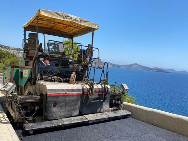 Pavimentadora asfáltica colocando una carretera costera. Cielo azul, mar y montañas de fondo.