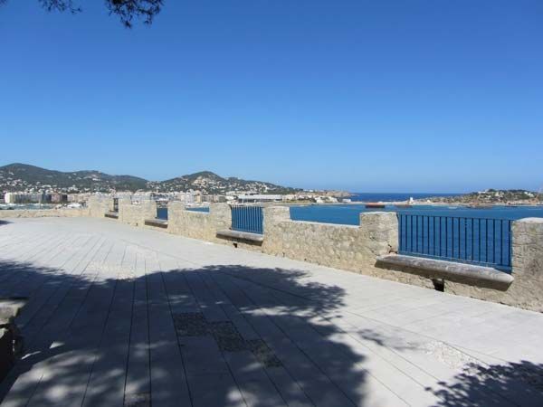 Mirador con vistas al mar con paredes de piedra, barandilla azul y un paisaje urbano a lo lejos bajo un cielo azul claro.