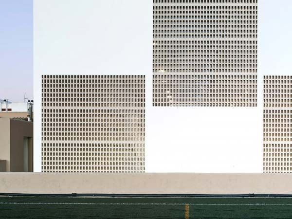 Fachada de edificio blanco con ventanas en forma de cuadrícula contra un cielo pálido, situado sobre un campo deportivo verde.