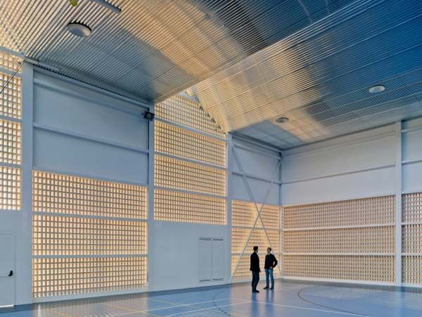 Dos personas se encuentran dentro de un gimnasio lleno de luz, con paredes de bloques de vidrio y un techo de metal.