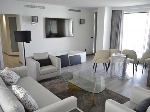 Sala de estar con muebles blancos, televisor y mesa de centro de cristal. Paredes gris claro y una ventana con cortinas.