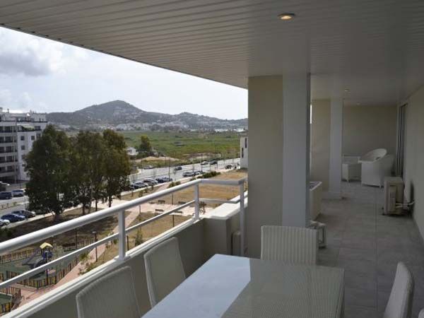 Vista desde el balcón de un paisaje urbano con una mesa blanca, sillas y colinas distantes bajo un cielo nublado.