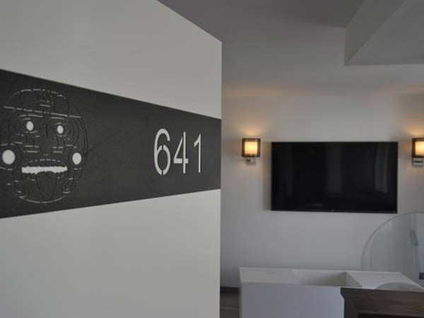 Placa con diseño del número de la habitación 641; vista interior de una habitación con TV, apliques y muebles blancos.