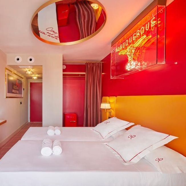 Habitación de hotel roja y amarilla con dos camas, un espejo y un letrero de neón en la pared.