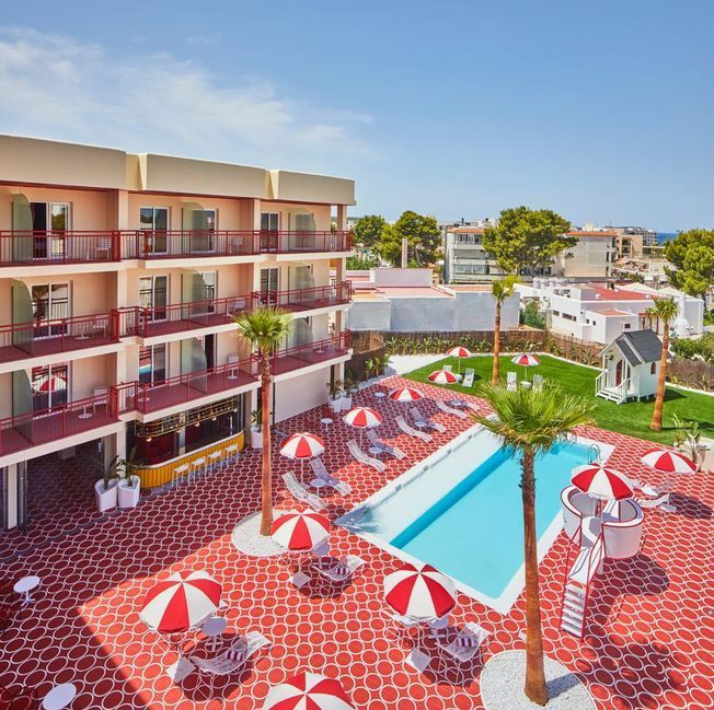 Vista de la piscina de un hotel con detalles en rojo y blanco, puesto de salvavidas, palmeras y tumbonas.