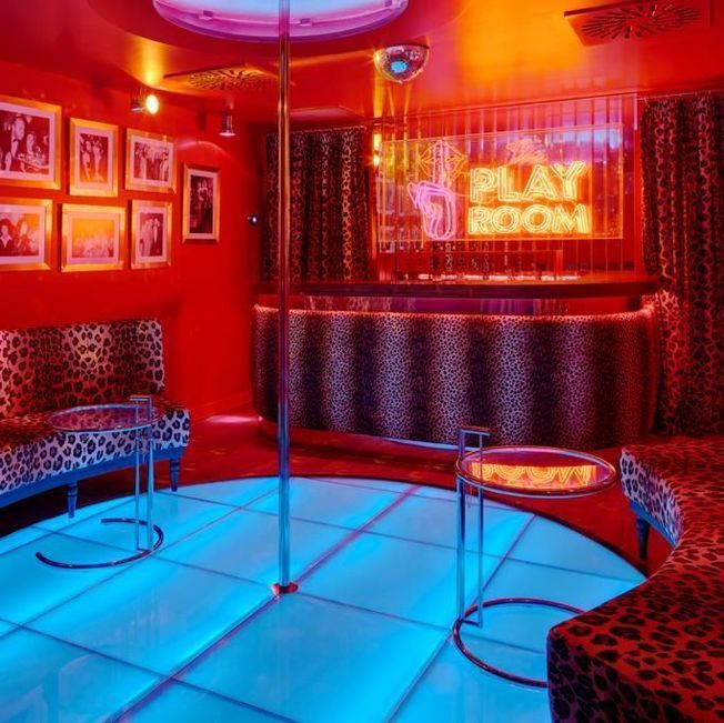 Interior de discoteca iluminado en rojo con pista de baile, barra, muebles con estampado de leopardo y un cartel de neón que dice